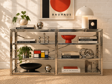 Load image into Gallery viewer, Image 1 of 23: A modern two-tier glass and metal shelving unit styled with books, decorative bowls, plants, and a lamp in a bright room with a white brick wall and framed Bauhaus art.
