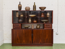 Load image into Gallery viewer, A mid-century modern rosewood hutch cabinet with open and closed compartments, styled with various ceramic vases and bowls, set against a white brick wall.