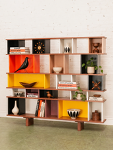 Load image into Gallery viewer, A mid-century modern wooden open bookcase shelf with multiple cubby compartments in warm wood, black, white, orange, and yellow, styled with books, vases, bowls, and a potted plant against a white brick wall.