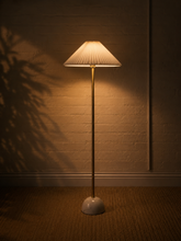 Load image into Gallery viewer, A tall floor lamp with a pleated beige shade and a slim gold-toned stand, glowing warmly against a textured brick wall and carpeted floor.