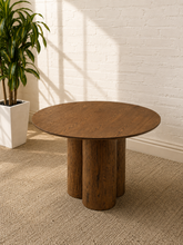 Load image into Gallery viewer, A round wooden side table with three cylindrical legs, shown in a styled room setting with a white brick wall, textured beige rug, and a potted green plant.