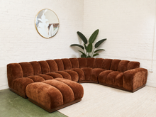 Load image into Gallery viewer, A curved modular sectional sofa upholstered in rich rust-brown velvet, set in a bright room with a white brick wall, a green rug, and a potted plant.