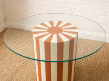 Load image into Gallery viewer, Round glass dining table with orange and cream sunburst striped cylindrical pedestal base.