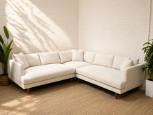 Load image into Gallery viewer, A modern L-shaped sectional sofa upholstered in off-white fabric with light wood legs, styled in a bright room with a white brick wall, beige rug, and potted plants.