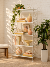 Load image into Gallery viewer, A tall white metal ladder-style bookshelf styled with books, vases, a plant, and decorative objects in a bright room with a white brick wall and potted green plants.