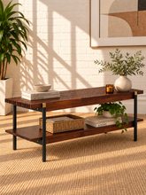 Load image into Gallery viewer, Image 2 of 5: A two-tier wooden console table with a dark brown finish and black metal legs, styled with books, a wicker basket, and potted plants in a bright room with warm sunlight and brick wall shadows.