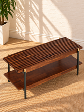 Load image into Gallery viewer, Image 4 of 5: A modern wooden coffee table with a dark brown slatted top, a lower wooden shelf, and black metal legs with brass accents, set on a woven jute rug against a white brick wall.