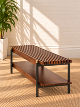Load image into Gallery viewer, Image 5 of 5: A modern wooden coffee table with a dark brown slatted top, a lower shelf, and a black metal frame with brass corner accents, set on a woven rug against a white brick wall.