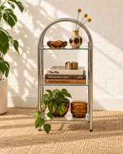 Load image into Gallery viewer, A three-tier glass and metal shelving unit styled with books, vases, and potted plants in a bright room with a white brick wall and natural sunlight.