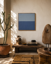 Load image into Gallery viewer, A minimalist blue and gray two-tone square wall art print in a thin natural wood frame, displayed in a rustic, sunlit room above a wooden bench with decorative pottery and a cactus.