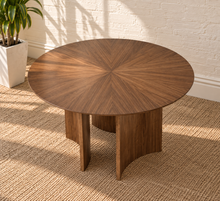 Load image into Gallery viewer, A round wooden coffee table with a sunburst grain pattern on the top and three curved block legs, set on a woven jute rug against a white brick wall.