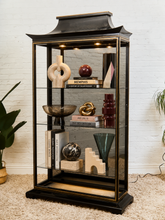 Load image into Gallery viewer, A tall black glass display cabinet with gold trim and a pagoda-style roof, styled with books and decorative objects on a beige carpet against a white brick wall.