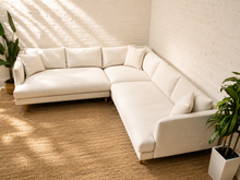 Load image into Gallery viewer, A modern L-shaped sectional sofa upholstered in off-white fabric, styled in a bright room with a white brick wall, a natural jute rug, and potted green plants.