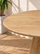 Load image into Gallery viewer, A close-up of a light natural wood dining table with a rounded edge and tapered pedestal base, set in a bright room with a white brick wall, woven rug, and potted plant.