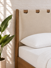 Load image into Gallery viewer, A close-up of a bed with a light beige fabric headboard featuring wooden eyelets, a white pillow, and a white sheet, set against a white brick wall with a potted green plant on the left.