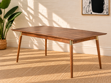 Load image into Gallery viewer, Image 1 of 4: A mid-century modern wooden dining table with tapered legs and brass metal accents, set in a room with a white brick wall, a potted plant, and a woven rug.