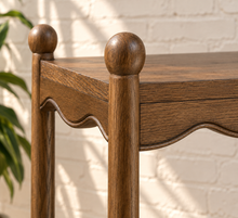 Load image into Gallery viewer, Image 5 of 5: Close-up of a wooden table corner with round turned posts and a scalloped apron, shown against a white painted brick wall with soft leaf shadows.