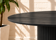 Load image into Gallery viewer, Image 2 of 4: A round black dining table with a fluted pedestal base, set on a woven beige rug against a white brick wall with warm sunlight and shadow patterns.