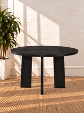 Load image into Gallery viewer, Image 2 of 4: A round black dining table with thick block legs, set on a woven jute rug against a white brick wall with sunlight casting shadows. A potted green plant is visible on the left.