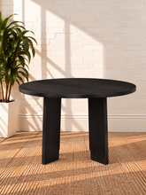 Load image into Gallery viewer, Image 3 of 4: A round black table with thick block legs, shown in a styled interior setting with a white brick wall, a potted plant, and a woven rug.