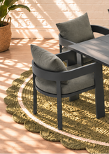 Load image into Gallery viewer, Image 2 of 4: Dark gray outdoor dining chairs with olive green cushions arranged around a matching dark gray table on a round braided jute rug, set in a sunlit patio area.