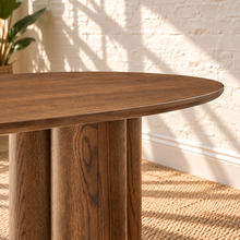 Load image into Gallery viewer, Close-up of a round wooden table with a medium brown finish and chunky cylindrical legs, set in a room with a white brick wall, woven rug, and soft window shadows.