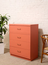 Load image into Gallery viewer, A four-drawer chest of drawers in a warm coral-orange color, styled in a room setting against a white brick wall with a green potted plant on the left and a wooden chair on the right.