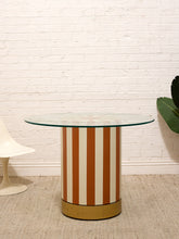 Load image into Gallery viewer, A mid-century modern cylindrical side table with vertical terracotta and cream stripes on a brass base, topped with a clear glass surface, set against a white brick wall.