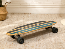 Load image into Gallery viewer, A longboard skateboard with a weathered wood deck featuring teal and black stripes, resting on a textured beige rug in a home setting.