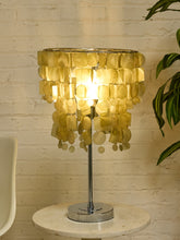 Load image into Gallery viewer, A table lamp with a circular shade made of overlapping translucent gold discs, mounted on a polished silver metal stand and base, set on a round marble side table against a white brick wall.