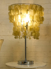 Load image into Gallery viewer, A table lamp with a circular shade made of overlapping translucent gold-toned discs, glowing from within on a polished silver stand against a light brick wall.