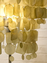 Load image into Gallery viewer, A close-up of a hanging decorative chandelier made of layered translucent oval and rectangular panels in warm gold and beige tones, with a softly glowing light behind it.