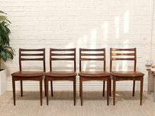 Load image into Gallery viewer, Four wooden dining chairs with brown leather seats arranged in a row against a white brick wall.