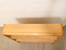 Load image into Gallery viewer, A light natural wood console table with a curved top and two open shelves, positioned against a white painted brick wall on a textured beige rug.