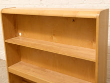 Load image into Gallery viewer, A light natural wood bookshelf with three open shelves, photographed against a white brick wall and carpeted floor.