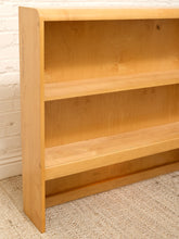 Load image into Gallery viewer, A light natural wood bookshelf with three open shelves, positioned against a white brick wall on a beige woven rug.