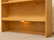 Load image into Gallery viewer, A close-up of a light natural wood bookshelf with three shelves, featuring a small under-shelf LED light glowing warmly, set against a white brick wall and carpeted floor.