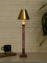 Load image into Gallery viewer, A table lamp with a warm brass-colored shade and base, a pink and brown marbled column, and a lit bulb, set on a tabletop against a white brick wall with a mug and plant nearby.
