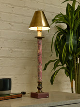 Load image into Gallery viewer, A table lamp with a warm brass-colored metal shade and base, featuring a slender pink marbled stem, shown lit on a tabletop next to a green potted plant against a white brick wall.