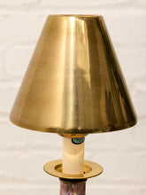 Load image into Gallery viewer, A close-up of a brass table lamp shade with a matching brass base ring and a cream-colored candle-style bulb, set against a white brick wall background.