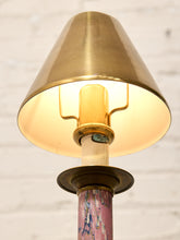Load image into Gallery viewer, A close-up of a table lamp with a warm brass-colored metal shade and base, a glowing light bulb, and a marbled pink, purple, and blue column, set against a white brick wall background.