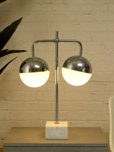 Load image into Gallery viewer, A modern two-light table lamp with polished chrome spherical shades and a white marble base, shown lit against a beige brick wall.