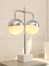 Load image into Gallery viewer, A modern two-light table lamp with polished chrome dome shades and a white marble base, shown lit against a white brick wall.