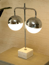 Load image into Gallery viewer, A modern two-light table lamp with polished chrome-finished spherical shades and glowing white diffusers, mounted on a square white marble base against a light brick wall.