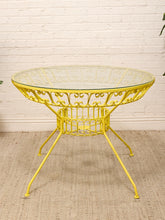 Load image into Gallery viewer, A bright yellow metal patio table with a round clear glass top, ornate scrollwork base, and four legs, set on a woven rug against a white brick wall.