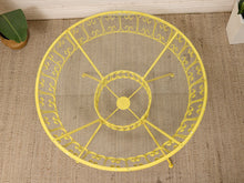 Load image into Gallery viewer, A bright yellow metal circular stand or frame with decorative scrollwork, photographed from above on a beige woven rug with small plant and wall decor visible at the edges.