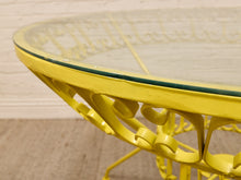 Load image into Gallery viewer, Close-up of a bright yellow metal table with a clear glass top, showing the decorative scrollwork frame and glass edge against a light wall and floor.