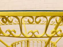 Load image into Gallery viewer, Yellow metal table frame with an ornate scrollwork design and a clear glass tabletop, shown against a white brick wall background.