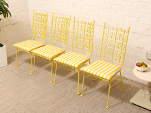 Load image into Gallery viewer, Four yellow metal dining chairs with ornate scrollwork backs and yellow-and-white striped upholstered seats, arranged in a row against a white brick wall.
