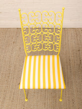Load image into Gallery viewer, A yellow metal side chair with an ornate scrollwork backrest and a yellow-and-white striped upholstered seat, shown on a woven rug against a white brick wall.
